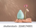 Small photo of Two daisy spring flowers near a tiny green vase on a rock on table. Misplaced eccentric. Fragile. assumptions. Unordinary. Brown background with copy space. Blooming flowers. Greeting card. sunlight