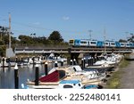 Small photo of Melbourne, Australia - January 20, 2023: View along Mordialloc Creek with moored fishing boats in a line and a suburban train crossing over the creek.