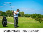 Small photo of Asian senior man holding golf club hitting golf ball on fairway at country club in sunny day. Healthy elderly male golfer enjoy outdoor activity sport golfing at golf course on summer vacation