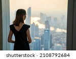 Asian woman standing by the window at skyscraper office building in metropolis and looking cityscape at summer sunset. Beautiful female relax and enjoy outdoor lifestyle activity in the city at night