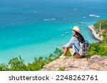 Asian woman with backpack travel at tropical island and resting on mountain peak in summer sunny day. Attractive girl enjoy outdoor lifestyle looking beautiful ocean nature on beach holiday vacation.