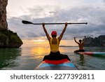 Asian people sitting on paddle board and rowing in the ocean together at tropical island at sunset. Man and woman enjoy outdoor lifestyle water sports surfing and paddle boarding on summer vacation.