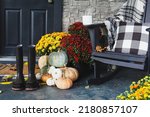 Small photo of Hot coffee sitting on rocking chair on front porch that has been decorated for autumn with white, orange, grey pumpkins, rain boots and mums. Selective focus with blurred foreground and background.
