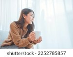 Happy asian woman relaxing drinking hot coffee or tea in holiday morning vacation on armchair at home, Cosy scene, Smiling pretty woman drinking hot tea in autumn winter. copy space.