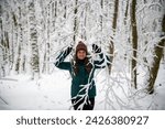 Captured within a snowy forest, a woman stands smiling, hands raised to the sky framed by the patterns of snow-laden branches. She is warmly dressed in a teal coat and a multicolored winter hat