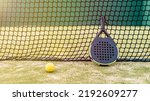Small photo of Padel racket and yellow ball behind net on a green court grass turf outdoors at sunset. Paddle is a racquet game. Professional sport concept with space for text