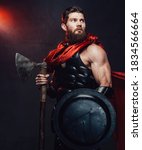 Small photo of Warlike and armoured rome empire fighter with beard and muscular build posing holding shield and axe in dark room with spotlight.