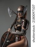 Small photo of Portrait of furious valkyrie dressed in armor holding huge axe and looking at camera against grey background.