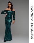 Small photo of Full-length portrait of a model-esque brunette with voluminous hairstyle in a graceful green evening gown on gray