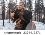 Small photo of A fierce Viking warrior poised with an axe and shield in a snowy pine forest, depicting strength and historical Nordic culture