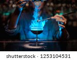 Bartender girl adding to a brown cocktail and pour on a flamed badian on tweezers a powdered sugar on the bar counter in the blue light.