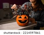 Small photo of hand of woman florist gently decorate lovely autumn arrangement of thistles flowers and plants in bright orange pumpkin with carved eyes and mouth. Beautiful floral decor for halloween. Close-up