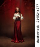 Small photo of Art photo with noise. Fairy gypsy woman medieval witch stands in dark room. Long black hair, rose hairpin. Red costume vintage dress, fortune teller costume Gold jewelry . Mystical fantasy girl
