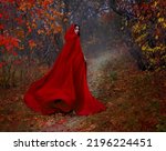 Small photo of Art gothic fantasy woman like red riding hood walks in dark autumn forest. long silk cloak flutters, waving fly in wind fabric in motion. Girl princess looks back. Fallen orange leaves dark trees