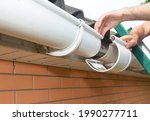 Small photo of Roof gutter installation and repair. A man on a ladder is replacing a plastic rain gutter joint, bracket, connecting the gutters together.