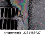 Small photo of An oil slick against the backdrop of an asphalt road flows into a storm drain through a grate. Environmental problems of water pollution.