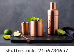 Small photo of Moscow mule cocktail in copper mug, shaker with fresh mint, lime on marble board. Dark background. Copy space.