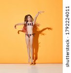 Small photo of Nice little girl with rainbow colored cornrows in striped bikini swimsuit jumping on place joyfully raising hands up. Full length shot isolated on orange, copy space. Childhood, snappy dresser, summer