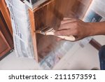 Small photo of A handyman paints a fresh coat of varnish on the surface of a base kitchen cabinet with a medium sized brush. Home renovation or finishing works.