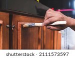 Small photo of A handyman paints a fresh coat of varnish on the surface of a base kitchen cabinet with a medium sized brush. Home renovation or finishing works.