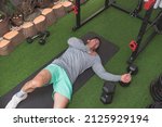 A man grimaces in pain after suffering a minor pectoral tear or chronic rotator cuff injury. Lying on a black mat at a home gym. Workout injury.