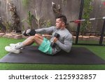 Small photo of Full Situps with Dumbbells and Alternate Cross Punch. Performed by a fit asian guy lying on a black mat. Intense reciprocal exercise at a home gym. SIde view.
