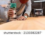 Small photo of A carpenter uses a wood trimmer to chamfer to edge of a desk. Compact Wood Palm Router Tool. At a furniture making workshop.