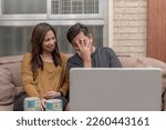 Small photo of A woman enthused at her husband covering his eyes while watching a horror movie on their laptop. Casual and lighthearted scene at the living room.