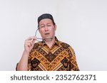 Small photo of A groggy and stressed middle aged man takes off his glasses after feeling eyestrain. An overworked person wearing a batik shirt and songkok skull cap. Isolated on a white background.