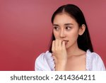 Small photo of An anxious and jittery young Asian woman biting her nail with a look of concern against a vibrant red background.
