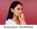 Small photo of An anxious and jittery young Asian woman biting her nail in an attempt to calm down. Portrait shot against a vibrant red background.