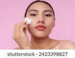Small photo of A pensive young Asian woman using a cotton ball to apply toner or astringent on her cheeks. Isolated on a pink background.