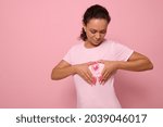 Small photo of Isolated portrait on colored background of mixed race woman in pink t-shirt, putting her hands on her chest in shape of heart with a pink satin ribbon in the center. World Cancer Awareness Day.