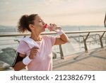 Small photo of Middle-aged female athlete in a pink T-shirt and gray leggings, with white wristbands, holds a terry towel on her shoulders and drinks water after workout standing on the city bridge at sunrise