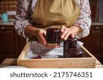 Cropped view of a woman in kitchen apron holding a glass jar of redcurrant jam over a wooden box with berry marmalade jars upside down. Collection of homemade preserves, confitures, jelly, canned food