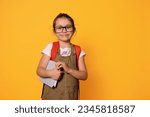 Small photo of Advertising studio portrait of Caucasian chappy smiling elementary age school girl with backpack, holding a textbook, dressed in casual clothes and eyeglasses, looking at camera, isolated background