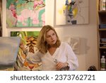 Authentic portrait of confident charming mature woman in white casual shirt, standing against a wall with displayed painted images, explaining a lesson of fine art at creative workshop or art gallery