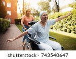 A woman with cancer is sitting in a wheelchair. She walks on the street with her daughter and they fool around. They are fun and they laugh. They walk in the courtyard of the clinic.