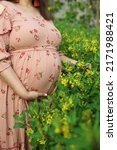 Small photo of A young woman in pink printed tight-fitting dress embraces her belly with a future baby with trepidation and tenderness. yellow flowers . selective focus" " follow focus" " shallow depth of field"
