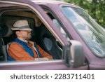 Small photo of A young male farmer in work clothes is resting in a car behind the wheel after work. A handsome brunette guy in a feller and a vest sleeps in the cab of his old car in the shade