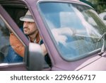 Small photo of A young male farmer in working clothes sits in a car behind the wheel. Handsome brunette guy in a feller and a vest driving his old car