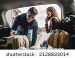 Young beautiful woman talking on the phone making a call from the back of her car sitting in the trunk while her boyfriend or husband it taking luggage baggage and other belongings from the car travel