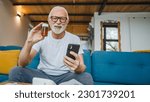 Small photo of One senior man sitting at home on sofa bed holding bottle of medicine pills medicament or supplement and smartphone read label instructions of drug tablet painkillers or vitamin ready to take medicine