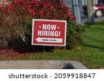 Small photo of Outdoor lawn sign now hiring apply inside with direction arrow, selective focus. Great resignation, employment, understaffed business, work strikes, absent workers due to covid labor shortage concept.