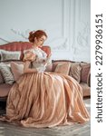 Small photo of Red-haired girl in a ball gown with a fan in her hands on a sofa in a bright room