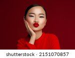 Sending a kiss with a blowing kiss. Fashion portrait of young asian model with red lips make up posing against red background.