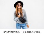 Fashionable portrait of smiling elegant brunette girl in sunglasses , black trendy hat and grey sweater posing over white wall. Bum bag. Positive mood.