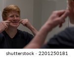 Sad caucasian redhead man in black t-shirt forces himself to smile by pulling his cheeks to the sides with his hands in front of mirror. Selective focus. Mental health theme.