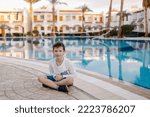 Small photo of A cheerful child in a long-sleeved T-shirt sits by the pool. Rest in the hotel. Happy holidays. Winter or autumn at the resort. Warm evening. swimming pool is heated. Free childhood. child lost a toot