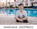 Small photo of A cheerful child in a long-sleeved T-shirt sits by the pool. Rest in the hotel. Happy holidays. Winter or autumn at the resort. Warm evening. swimming pool is heated. Free childhood. child lost a toot